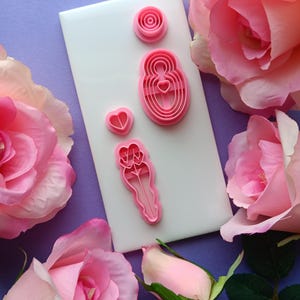 May include: Pink plastic cookie cutters in various shapes, including a rose, a heart, and a figure, are arranged on a white tile. The tile is surrounded by pink roses against a purple background.