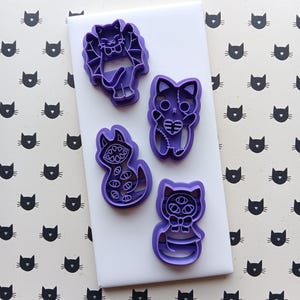 May include: A collection of five purple cat-shaped cookie cutters. The cutters feature various designs, including a cat with bat wings, a cat with a stitched heart, and a cat with multiple eyes. The cutters are on a white surface with a repeating black cat face pattern.