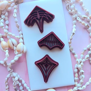 May include: Three red plastic cookie cutters on a white background. The cutters are shaped like a palm tree, a fan, and a diamond.