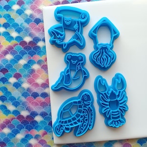 May include: A collection of five blue plastic cookie cutters in the shapes of sea creatures: a hammerhead shark, squid, frog, turtle, and lobster. The cutters are arranged on a white surface, with a colorful, patterned background.