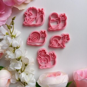 May include: Five light pink plastic cookie cutters shaped like snails, each featuring a unique shell design including a flower, a wheel, and a spiral pattern, arranged in a cluster alongside several artificial pink and white flowers.