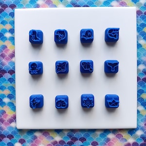May include: Twelve blue square stamps with various sea creature and nautical designs, including a whale, octopus, and ship's wheel, are arranged on a white surface. The stamps are set against a colorful, patterned background.