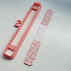 May include: A pink nail file with a white and pink floral pattern. The file is in a pink plastic case with a bow on the top.