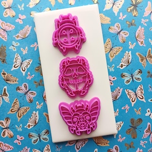 May include: Three pink cookie cutters in the shapes of a skull, a butterfly, and a decorative design. The cutters are on a white surface, set against a blue background with butterfly patterns.
