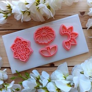 May include: Four pink flower-shaped cookie cutters on a white rectangular surface. The cutters are in various floral designs, including a daisy, a tulip, and a stylized flower. White flowers surround the cutters.