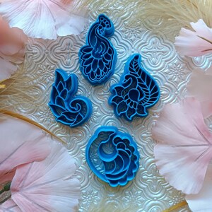 May include: Four blue floral-shaped cookie cutters. The cutters have intricate designs and are arranged on a textured, patterned surface. The background includes pink and white fabric flowers and dried grasses, creating a decorative display.