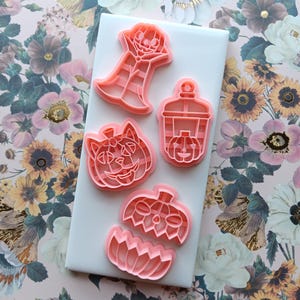 May include: Pink plastic Halloween-themed cookie cutters in various shapes, including a jack-o'-lantern, a ghost, and a lantern, arranged on a white surface. The cutters are set against a floral patterned background.