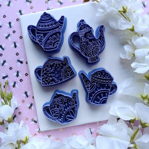 May include: Five blue teapot-shaped objects with intricate white patterns are arranged on a white surface. The teapots are surrounded by white flowers and set against a pink background with bee illustrations.