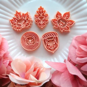 May include: Six orange plastic cookie cutters in various autumn-themed shapes, including leaves, acorns, and woodland animal faces. The cutters are arranged on a white plate, with pink flowers in the foreground.