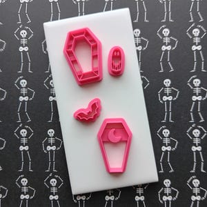 May include: Pink Halloween-themed cookie cutters in the shapes of a coffin, ghost, bat, and coffin with a crescent moon. The cutters are on a white surface, with a black background featuring a repeating skeleton pattern.