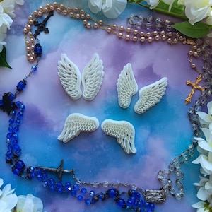 May include: Four white angel wings with a detailed feathered design, arranged in a cross shape on a blue and purple background. Two rosaries with blue and clear beads and silver and gold crosses are also visible.