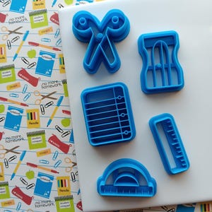 May include: A collection of blue plastic school-themed cookie cutters. The set includes scissors, a pencil sharpener, a notepad, a ruler, and a protractor. The cutters are arranged on a white surface with a colorful school-themed background.