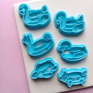 May include: Six light blue plastic cookie cutters in various animal shapes, including a duck, snail, and pig. The cutters are arranged on a white surface against a pink polka dot background. These are likely for baking.