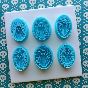 May include: Six light blue, oval-shaped cookie cutters with ghost designs. Each cutter features a different Halloween-themed scene, including a haunted house, stars, a moon, and flowers. The cutters are arranged on a white surface.