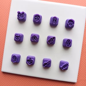 May include: A collection of twelve small, square, purple stamps with Halloween-themed designs. The stamps feature images of a ghost face, a bat, a pentagram, a coffin, and other spooky icons. They are arranged on a white square surface.