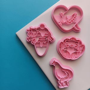 May include: A set of four pink plastic cookie cutters featuring various shapes: a heart with a snake, a cow on an ice cream cone, a pig surrounded by petals, and a chicken. Each cutter has a raised edge for cutting and an inner design.