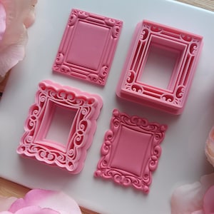 May include: Four pink, ornate frame-shaped cookie cutters. Each cutter has a different decorative design, including scrollwork and detailed edges. The cutters are arranged on a white surface, with pink flowers in the background.