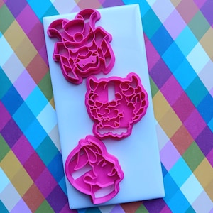 May include: Three pink plastic cookie cutters in the shapes of a samurai, a tiger, and a rabbit. The cookie cutters are arranged on a white rectangular surface, set against a colorful plaid background.