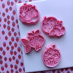 May include: Pink plastic cookie cutters in the shapes of a strawberry, a goldfish, and other strawberry-themed designs. The cutters are on a white surface with a pink strawberry pattern in the background.