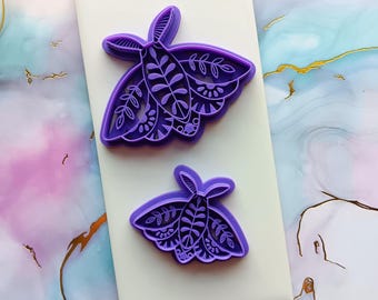 May include: Two purple plastic cookie cutters shaped like moths with intricate floral designs.