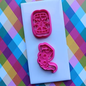 May include: Two pink plastic cookie cutters on a white surface. The top cutter is a rectangular shape with a starburst design. The bottom cutter is a stylized octopus. The background is a colorful plaid pattern.