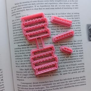 May include: Pink plastic dream symbol cookie cutters on a book page with the title "Symbolism of Dreams". The cutters are in various shapes, including a tiered design and rectangular shapes. The book is open to a page with text.
