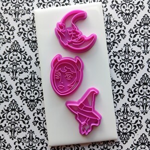 May include: Three pink cookie cutters in the shapes of a crescent moon with a face, a devil face, and a witch hat. The cutters are arranged on a white rectangular surface against a black and white patterned background.