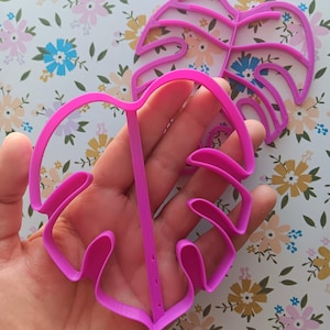 May include: Two pink plastic cookie cutters shaped like monstera leaves. The cutters are held in a hand.