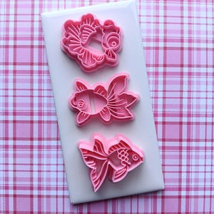 May include: Three pink plastic cookie cutters in the shape of goldfish on a white rectangular surface. The cookie cutters are arranged vertically, each with a different goldfish design. The background is a pink and white plaid pattern.