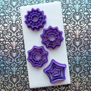May include: Four purple spiderweb-shaped cookie cutters on a white surface. The cutters are in various shapes, including round and star-like designs. The background features a patterned design with spiders and skulls.
