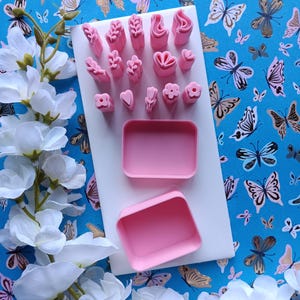 May include: A collection of pink polymer clay stamps in various floral and leaf designs, along with two pink rectangular containers, arranged on a white surface. The background features a blue paper with butterfly patterns. White flowers are in the foreground.