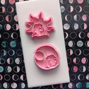 May include: Two pink plastic cookie cutters featuring stylized character faces. One cutter is shaped like a sun with radiating points, and the other is shaped like a crescent moon. Both cutters have internal details to imprint facial features.