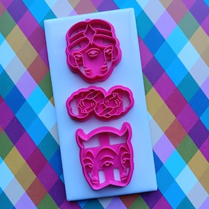 May include: Three pink plastic cookie cutters in the shapes of faces and a pair of roses are arranged on a white rectangular surface. The cookie cutters are on a colorful, patterned background.