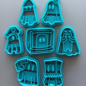 May include: Set of seven teal blue plastic cookie cutters shaped like ghosts wearing different costumes. The costumes include a pirate, a TV, a cat, a unicorn, and a few other variations.