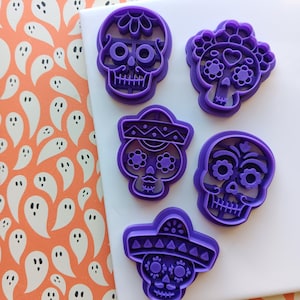 May include: Five purple skull-shaped cookie cutters with various floral and sombrero designs. The cutters are arranged on a white surface, set against a background of orange and white ghost-patterned paper. Ideal for Halloween or Day of the Dead baking.