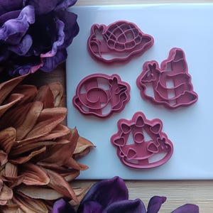 May include: Pink plastic cookie cutters in the shapes of a snail, a ram, a giraffe, and a cow. The cutters are on a white surface, with purple and brown floral decorations in the background.