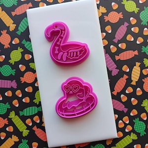 May include: Two pink plastic cookie cutters. One is a flamingo with bone details, the other is a ghost wearing heart-shaped glasses. The cutters are on a white surface, with a candy-themed background.