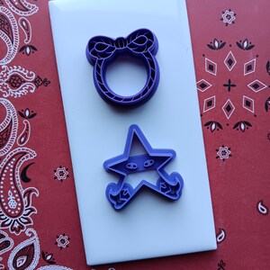 May include: Two purple cookie cutters on a white surface. One is a bow-shaped ring, and the other is a star with eyes and feet. The background is a red bandana with white paisley patterns.