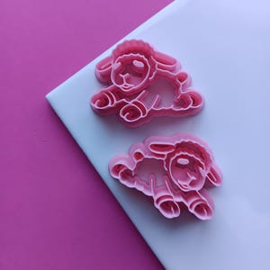 May include: Two pink plastic lamb-shaped cookie cutters. The cutters are lying on a white surface, with a pink background. The lamb cutters are detailed with eyes, ears, and curly wool. Ideal for baking themed cookies.