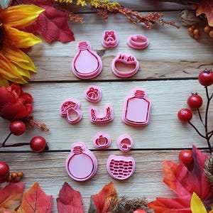 May include: A collection of pink cookie cutters in various shapes, including light bulbs, mushrooms, and other fall-themed designs. The cutters are arranged on a wooden surface, surrounded by autumn leaves and decorative elements.