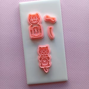 May include: Pink plastic cat-shaped cookie cutters on a white surface. The cutters include a cat with a clock face, a bow tie, and other cat-related shapes. The background is a light pink dotted pattern.