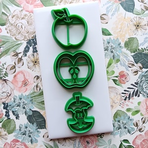 May include: Three green plastic apple-shaped cookie cutters. The cutters are in the shape of a whole apple, a half-eaten apple, and an apple core. The cutters are on a white surface, with a floral patterned background.