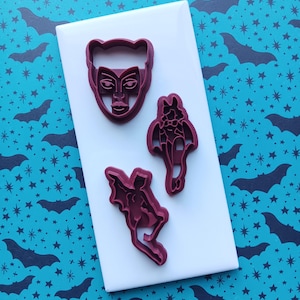 May include: Three dark red cookie cutters in the shapes of a masked face, a bat-winged figure, and a bat-winged figure in a flying pose. The cutters are on a white rectangular surface, set against a teal background with a pattern of bats and stars.