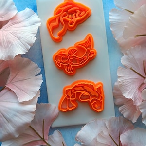 May include: Three orange koi fish cookie cutters on a white tile, set against a backdrop of pink and white flower petals and a blue surface. The cookie cutters are in different poses, showcasing the koi fish design.