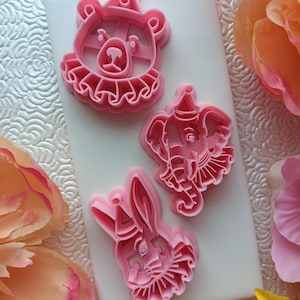 May include: Three pink plastic cookie cutters in the shapes of circus animals: a bear, an elephant, and a rabbit. Each animal wears a party hat and a ruffled collar. The cutters are on a white surface, surrounded by pink and peach flowers.