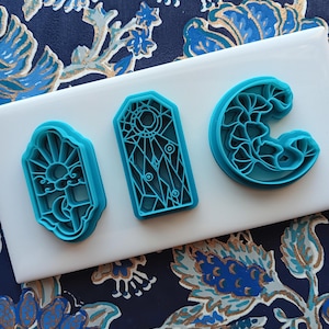 May include: Three turquoise cookie cutters in different shapes: a sun and moon, a stained glass window, and a floral crescent. The cutters are on a white surface against a blue floral patterned background.