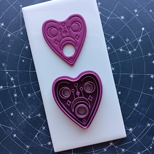 May include: A purple heart-shaped cookie cutter with a design of a Ouija board inside. The cutter is on a white surface.