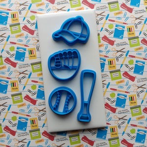 May include: Blue plastic baseball-themed cookie cutters on a white surface. The set includes a baseball cap, baseball glove, baseball, and bat. The background features a patterned fabric with school-related elements.