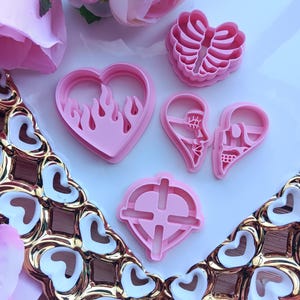 May include: A collection of pink heart-shaped cookie cutters in various designs, including flames, a rib cage, and a target. The cutters are arranged on a white surface with decorative gold and white heart-shaped accents and pink flowers.