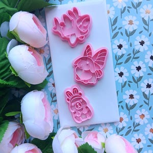 May include: Three pink cookie cutters in the shapes of bunnies and a bear on a white surface. The cookie cutters are arranged on a white surface, surrounded by pink and white flowers and a blue floral patterned background.